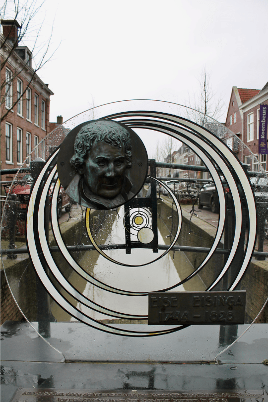 Monument ter ere van Eise Eisinga in Franeker, met een bronzen portret van de astronoom en een abstracte voorstelling van een planetarium, geplaatst boven een gracht met historische panden op de achtergrond.