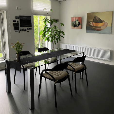 Een moderne eetruimte met een glazen tafel en vier stijlvolle stoelen. De ruimte is sfeervol ingericht met schilderijen aan de muur, een grote kamerplant en decoratieve elementen.