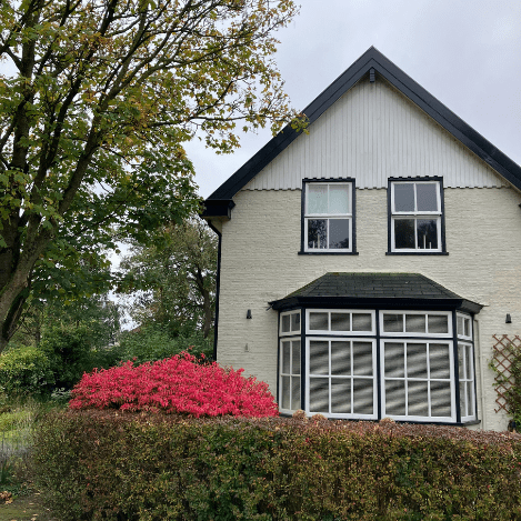 De voorgevel van een sfeervol wit huis met een erker, omgeven door een groene haag en kleurrijke bloemen.